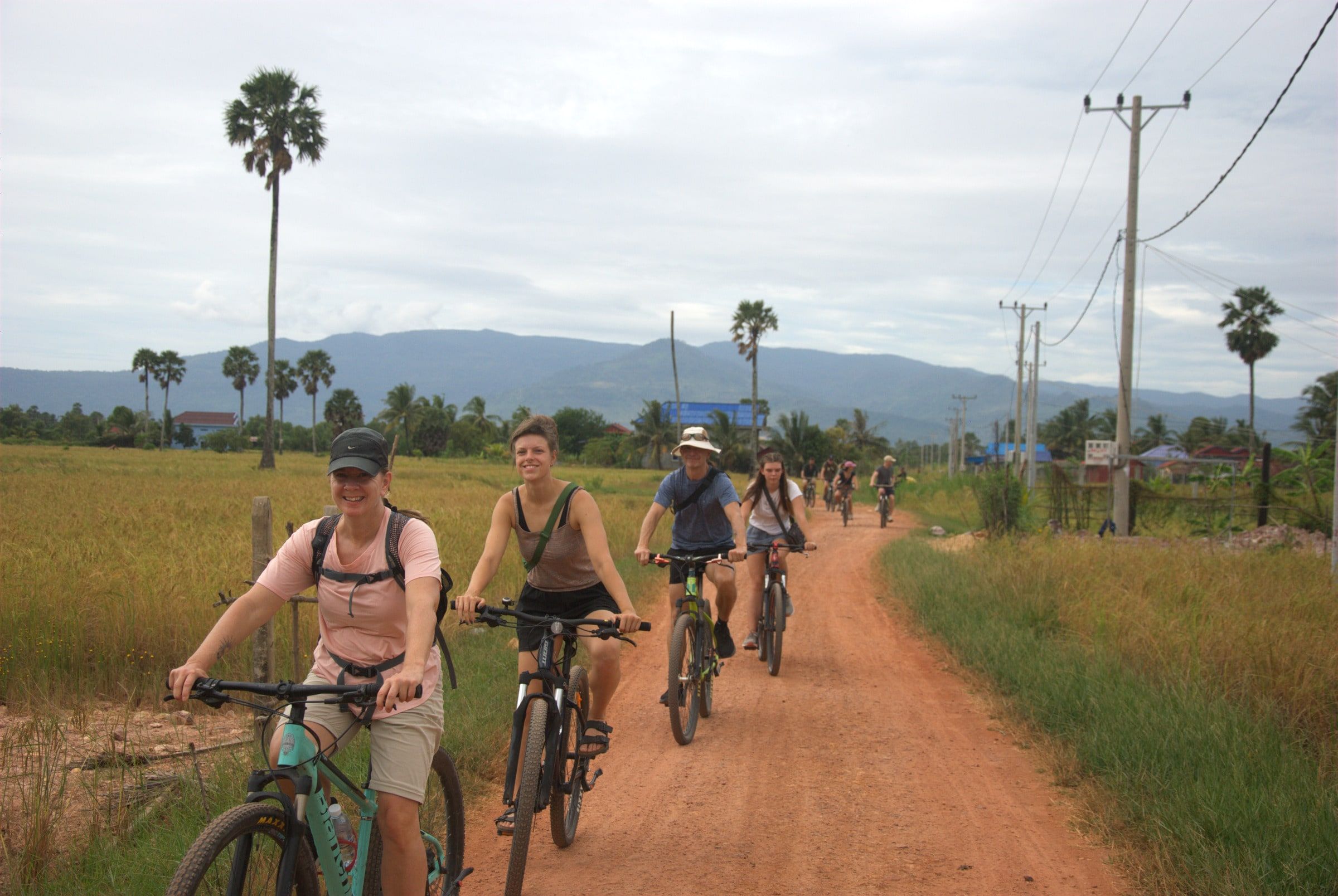 Guests riding bicycles down a red-dirt road between rice fields, mountains in the distance