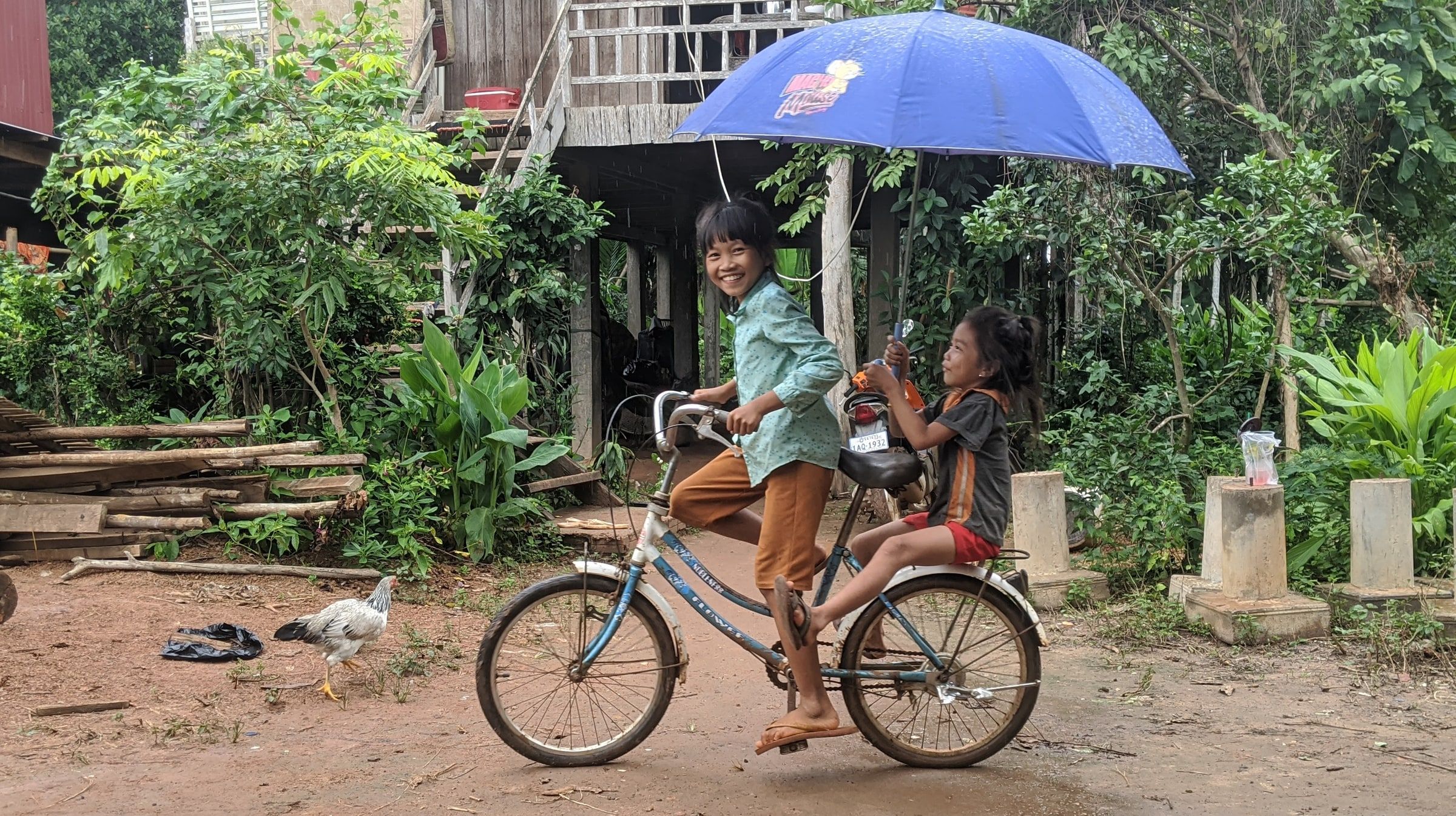 Two children riding one bicycle, the passenger holding an umbrella for shade