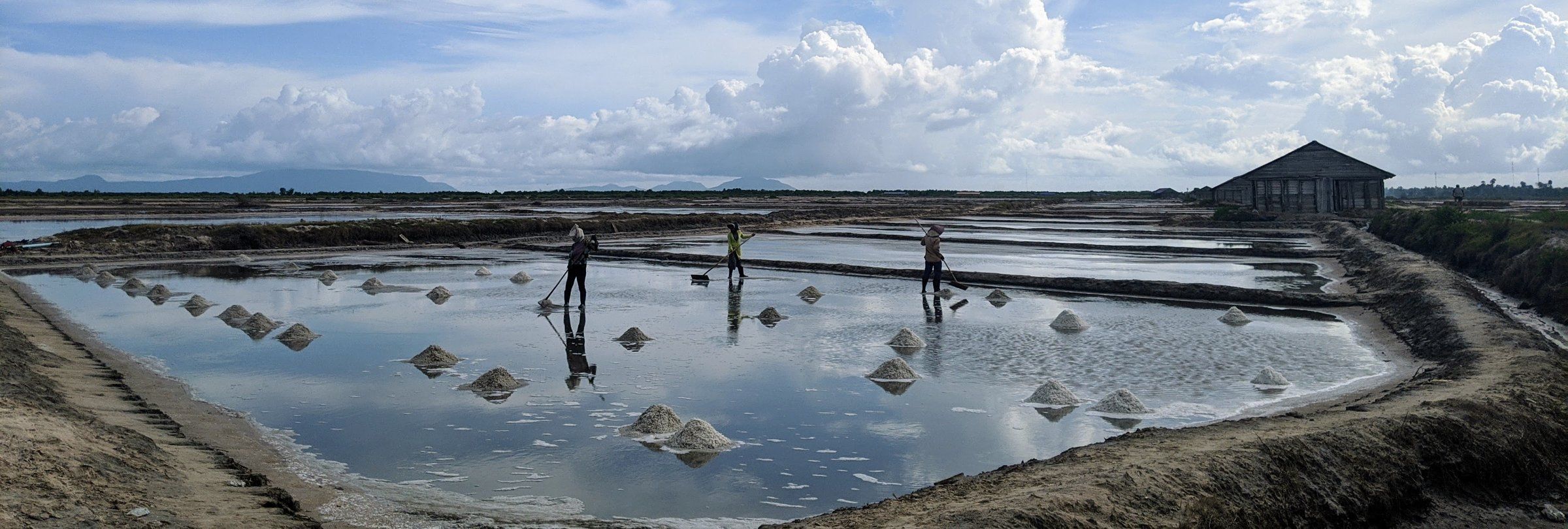 Early light across the shallow evaporation pools of the Kampot salt fields, a wooden salt barn with a rust-red roof behind