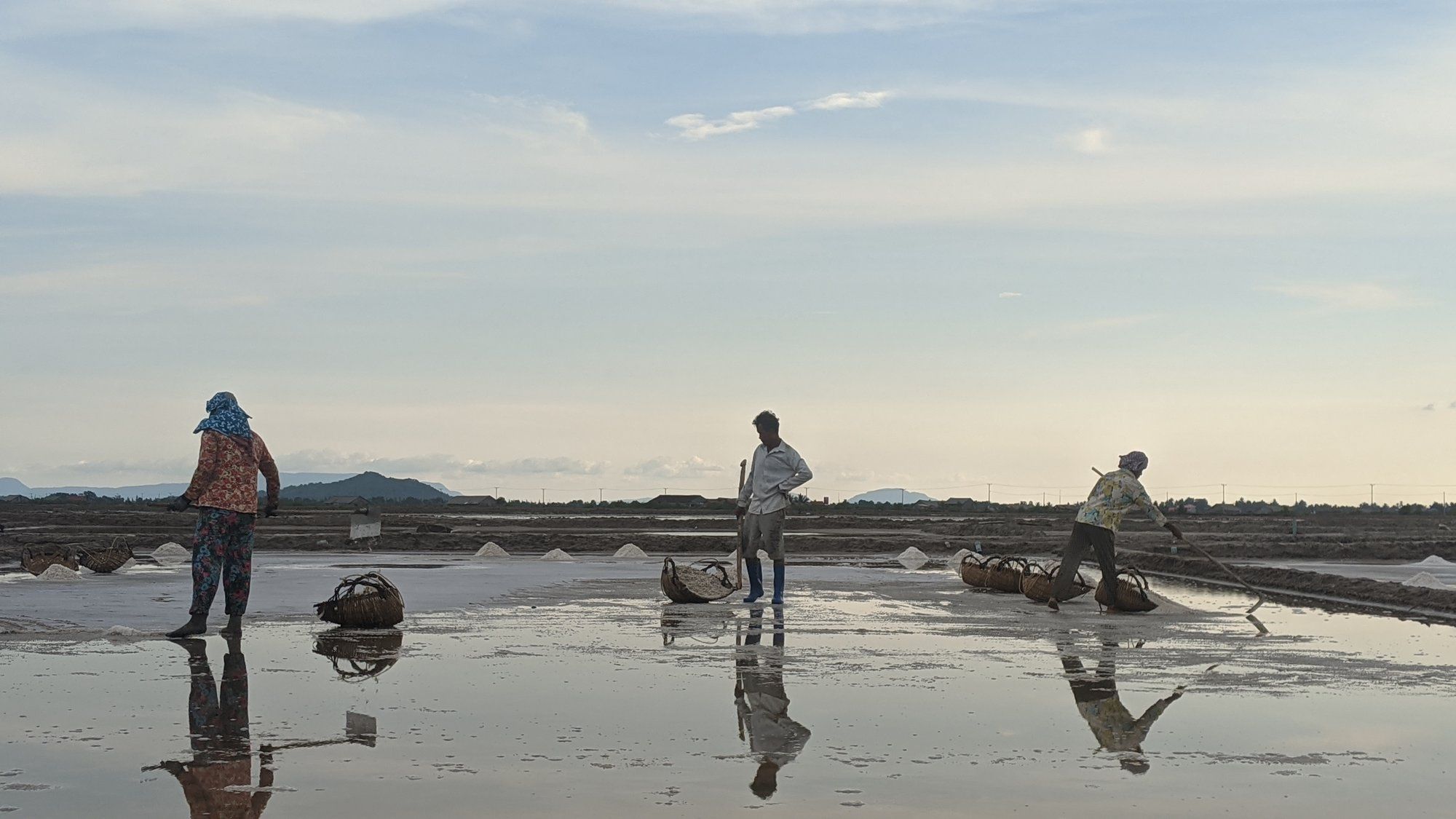 Three salt workers raking the pans at golden hour, reflected in the water